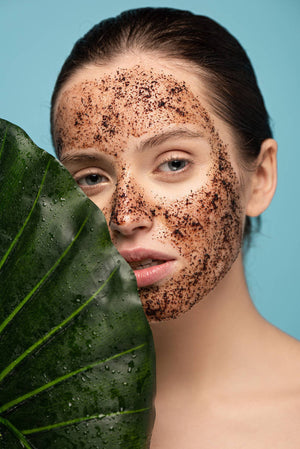 Woman with coffee scrub on her face next to a large green leaf.