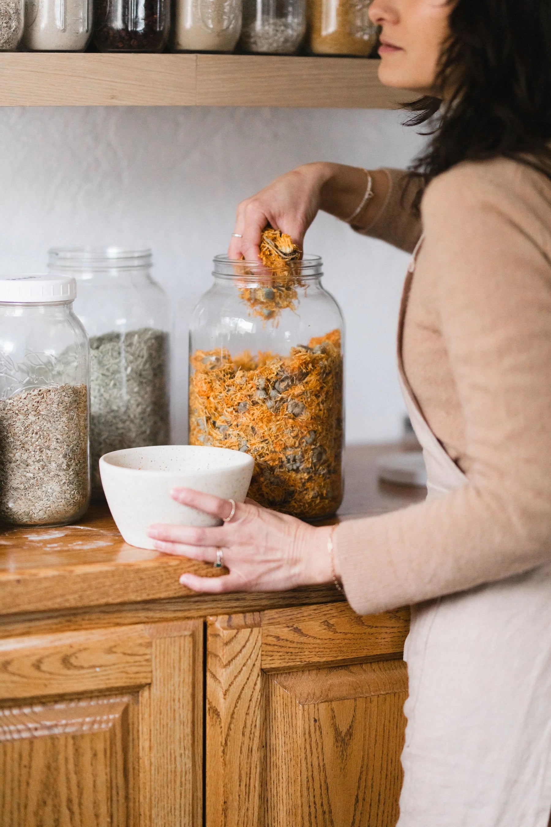 Person pouring dried herbs from a jar into a bowl on a wooden shelf.